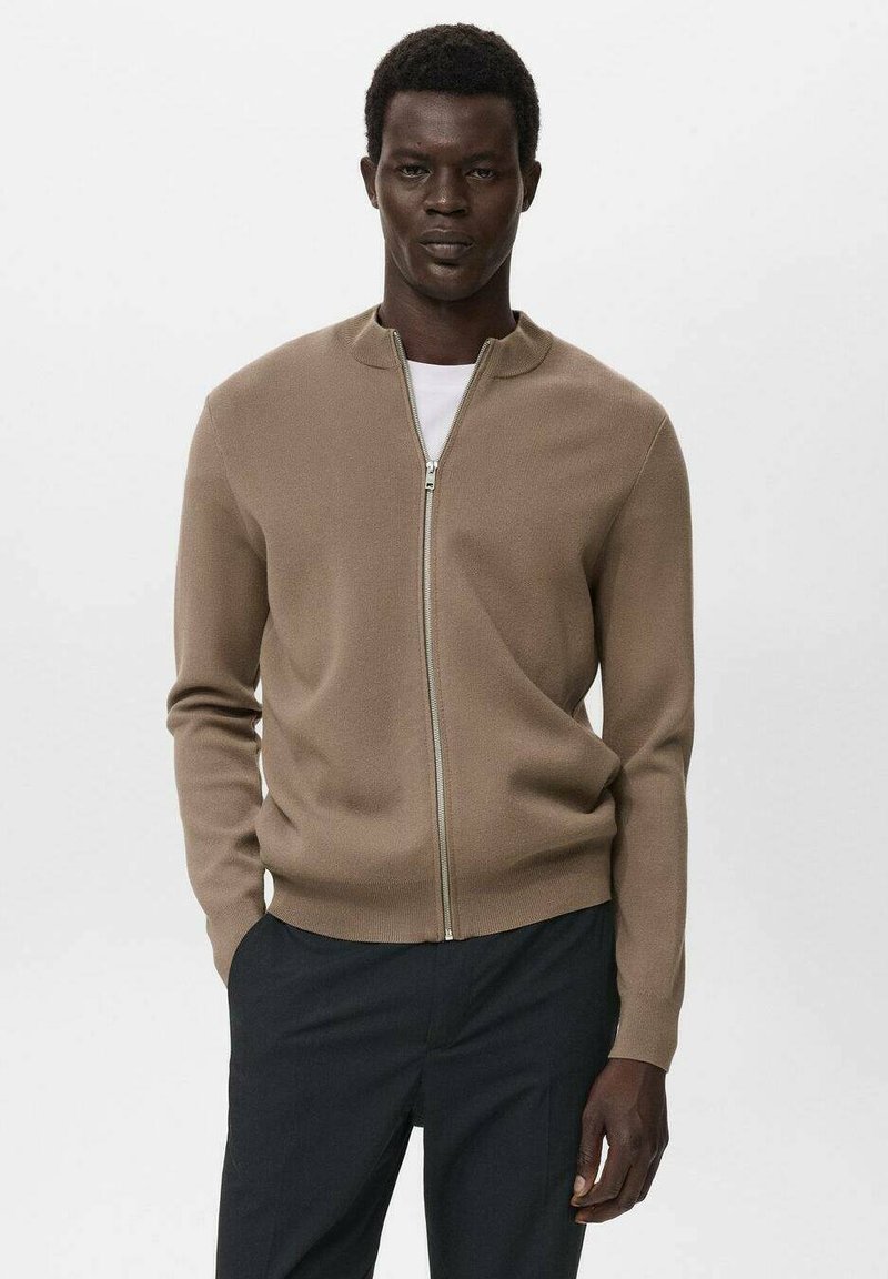 Man in een beige sweatshirt met rits over een wit shirt en donkere broek, staand met één hand in de zak tegen een effen achtergrond.