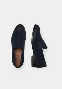 Marineblaue Wildleder Slipper mit runder Zehenform, genähten Details und einem dekorativen Riemen über der Vorderseite. Glatte schwarze Sohle.