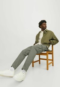 Chaqueta bomber verde oliva, camisa crema y pantalones cargo verdes oliva. El modelo lleva zapatillas blancas con suelas texturizadas, sentado en una silla de madera.