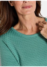 Frau mit langen braunen Haaren, die einen strukturierten grünen Pullover trägt, den Ausschnitt nahe dem Schlüsselbein haltend, Lippen leicht lächelnd und glänzend.