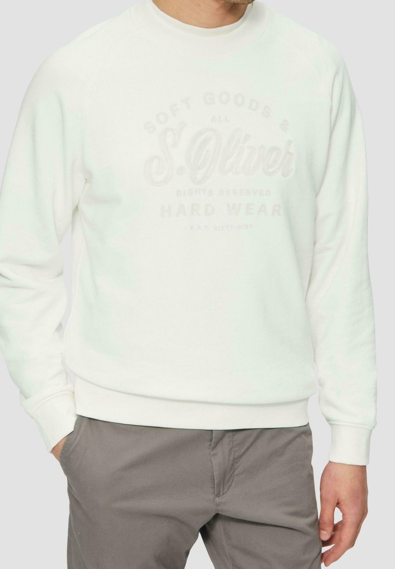Homme portant un sweat-shirt blanc avec un texte pâle et un pantalon gris, debout avec une main dans la poche sur un fond clair.