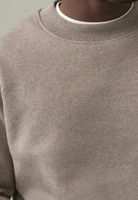 Sudadera de color marrón claro, con textura, que presenta un cuello redondo acanalado. La tela parece suave y ligeramente jaspeada, con un diseño minimalista.