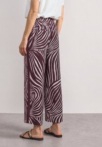 Personne portant un pantalon à jambes larges avec des rayures zèbre abstraites violettes et blanches et des sandales noires avec des brides dorées, debout sur un sol gris.