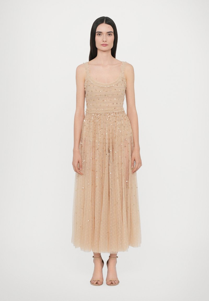 Abito in tulle beige con un corpetto aderente, spalline arricciate e una gonna fluida alla caviglia impreziosita da paillettes. Semplici sandali con tacco.