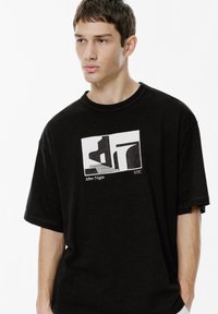 Ung mand iført en oversized sort t-shirt med en hvid grafik af trapper og arkitektoniske former, teksten lyder "After Night NYC."