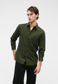 Σκούρο πράσινο πουκάμισο με κουμπιά σε υφασμάτινη υφή, διαθέτοντας τσέπη στο στήθος και διακριτό λογότυπο στο στρίφωμα, σε συνδυασμό με σκούρους τζιν παντελονιού.
