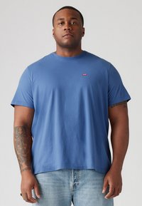 Rövid ujjú kék pamut póló, kerek nyakkivágással és kis piros Levi's logóval a bal mellen. A modellen világoskék farmernadrág is van.