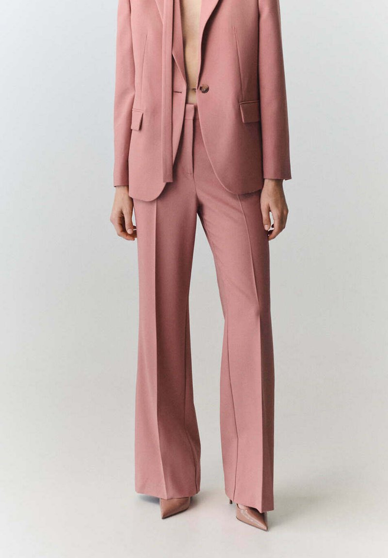 Femme portant un costume rose tailleur avec un pantalon à jambes larges et des escarpins pointus assortis sur un fond uni.
