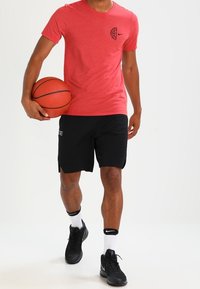 Osoba ubrana w czerwoną koszulkę i czarne szorty, trzymająca piłkę do koszykówki na wysokości bioder, stojąca na białym tle.