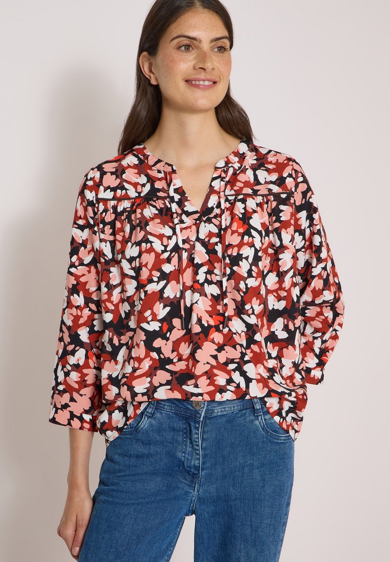 Femme portant un chemisier ample à motifs floraux rouges, roses, noirs et blancs, associé à un jean bleu, souriante et posant avec une main dans la poche.