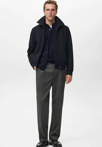 Chaqueta de cremallera azul marino con cuello alto, combinada con una camisa azul oscuro de botones y unos pantalones grises a medida. Unos zapatos negros completan el look.