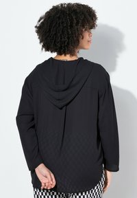 Schwarzes Kapuzenshirt mit langen Ärmeln, ausgestattet mit einem karierten Muster am hinteren Saum. Leichter Stoff, lockere Passform.