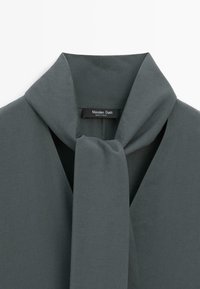 Blusa grigia con colletto drappeggiato e dettaglio a cravatta. Tessuto morbido con una consistenza setosa. Etichetta con la scritta "Massimo Dutti, made in Turkey."