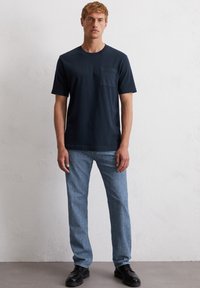 Marineblauwe katoenen T-shirt met een borstzak, gecombineerd met lichtblauwe denim broek en zwarte schoenen, tegen een neutrale achtergrond.