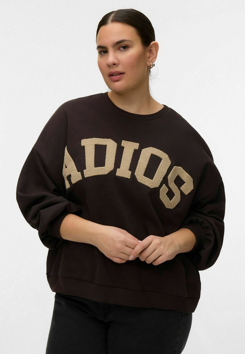 Femme aux cheveux foncés attachés, portant un sweat-shirt noir surdimensionné avec le mot « ADIOS » en grandes lettres beiges, les mains lâchement jointes.