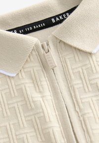 Beige zip-up poloing póló, textúrált szövött mintával, gallérral és fehér részletekkel, márkás címke látható a nyakvonal belsejében.