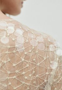 Tissu beige transparent avec un motif géométrique, agrémenté de sequins ronds et d'une broderie complexe. Texturé doux avec une apparence délicate.