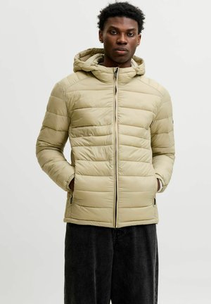 Jeune homme portant une veste matelassée à capuche beige, les mains dans les poches, debout devant un fond clair uni.