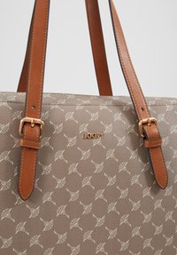 Beige mønstret håndtaske med lysebrune læderremme og guldspænder, der har små gentagne blomster motiver og brandnavnet "JOOP!" i guld.