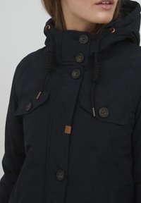 Marineblauwe jas met hoge kraag, knoopsluiting en trekkoorden. Voorzien van twee voorzakken en een klein logo-opdruk. Gestructureerde stof.