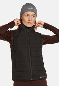 Black insulated vest with a high collar, zip closure, and side pockets. Worn over a brown long-sleeve top. Features a smooth, quilted texture.