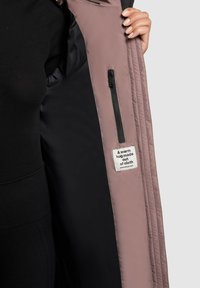 Chaqueta acolchada rosa con interior negro, que presenta un bolsillo con cremallera y una etiqueta de tela que dice "Un abrazo cálido hecho de tela."