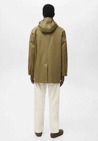 Persona de espaldas vistiendo una chaqueta con capucha de color marrón claro, pantalones blancos y zapatos marrones, contra un fondo blanco plano.