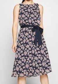 Květinové šaty s námořnickým pozadím, zdobené růžovými a bílými květinovými vzory, bez rukávů, s látkovým opaskem svázaným v pase.