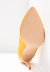 Chaussure à talon haut en daim jaune à bout pointu, inclinée avec la semelle tournée vers l'avant sur un fond blanc.
