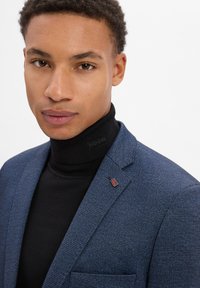 Marineblau strukturierter Blazer über einem schwarzen Rollkragenpullover, versehen mit einer kleinen Union Jack-Nadel am Revers. Glatter Stoff mit einer taillierten Passform.