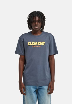 Jeune personne aux dreadlocks mi-longs portant un t-shirt bleu foncé avec le texte "ELEMENT WORLDWIDE" et un jean bleu clair sur un fond uni.