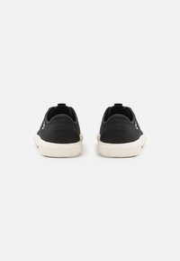 Zapatillas de lona negras con una superficie texturizada, suela exterior de goma blanca y acentos sutiles del logo en los lados. Diseño de perfil bajo con una lengüeta trasera.