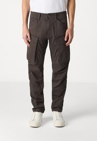 Donkerbruine cargobroek met meerdere zakken, ritsdetails en een taps toelopend ontwerp. G комбинированы met witte sneakers en een eenvoudige witte shirt.