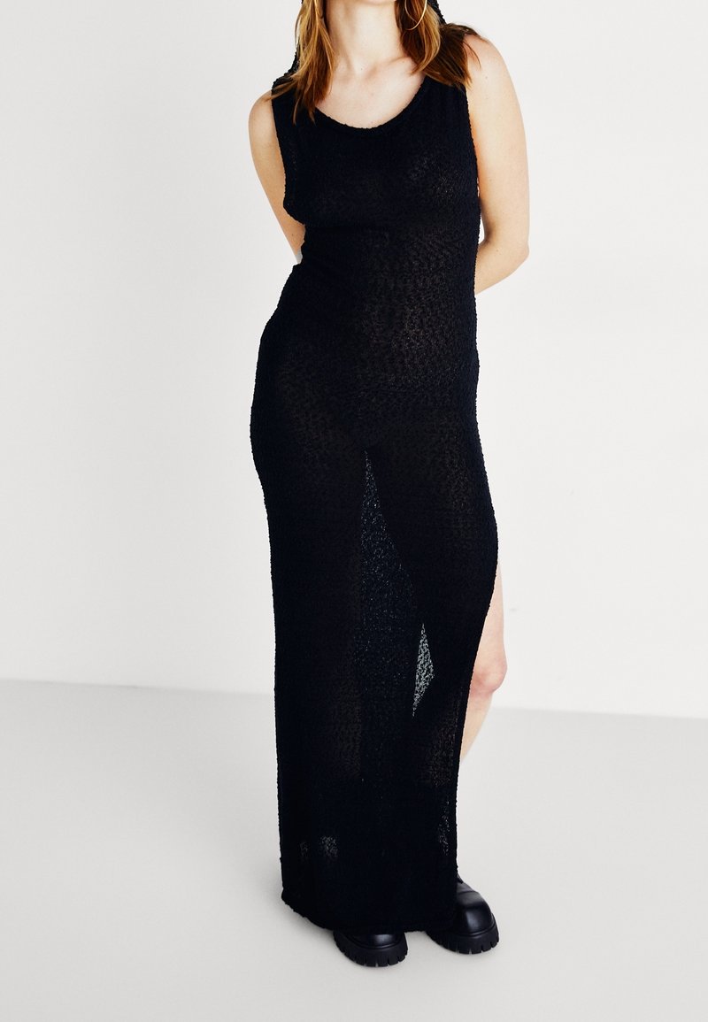 Mujer con un vestido maxi negro sin mangas y transparente, con una abertura lateral y zapatos negros gruesos, posando frente a un fondo blanco.