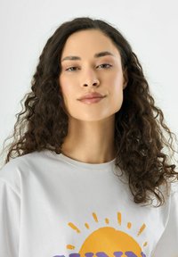 Weißes Baumwoll-T-Shirt mit einem großen orangen Sonnen-Graphic und lila Text darunter. Das Modell hat lange, lockige dunkle Haare.