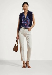 Mujer vistiendo una blusa azul con estampado, un chaleco de punto azul marino, pantalones blancos de cintura alta, tacones marrones y cargando un bolso marrón.