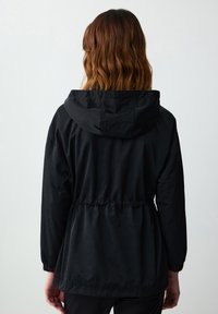 Zwarte lichtgewicht jas met een capuchon, cinch taille, elastische manchetten en een gladde textuur. Achteraan is het minimalistische ontwerp en de losse pasvorm zichtbaar.
