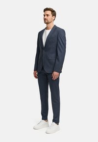 Marineblauwe structuurkostuum met een aansluitende blazer en op maat gemaakte broeken, gecombineerd met witte sneakers. Voorzien van een enkele knop en zijzakken.