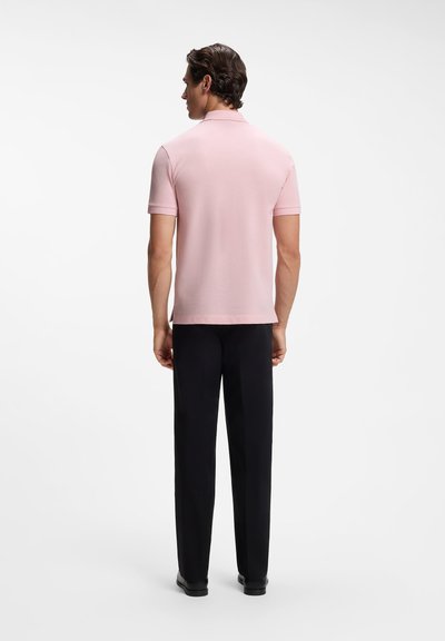 Homme de dos portant un polo manches courtes rose clair, un pantalon noir et des chaussures noires sur un fond blanc uni.