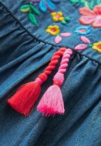 Tissu en denim avec broderie florale colorée et deux pompons tressés rouges et roses suspendus à la couture froncée.