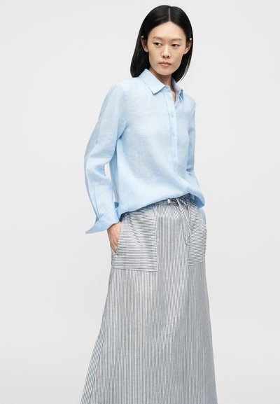 Kvinne med langt svart hår iført en lyseblå skjorte med knapper og et stripete langt skjørt med høy midje og store framlommer.