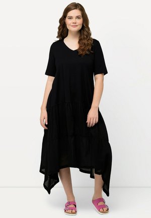 Schwarzes Kurzarmkleid mit V-Ausschnitt, gestuftem Saum und High-Low-Design. Getragen mit rosa Sandalen, die einen lässigen, fließenden Stil zeigen.