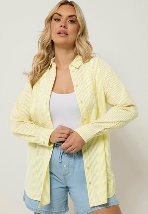 Lyshåret kvinde iført en lysegul, stribet skjorte med knapper over en hvid tanktop og lyseblå, højtaljede denimshorts.
