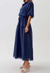 Marinblå, veckt maxiklänning i en texturerad tyg, korta ärmar och en knapp-up krage, kombinerad med ljusa, remförsedda klackskor.