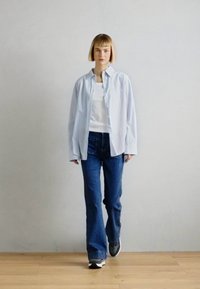 Camisa a rayas azul claro sobre una blusa blanca, combinada con jeans de campana azul oscuro y zapatillas grises. Texturas suaves, ajuste casual.