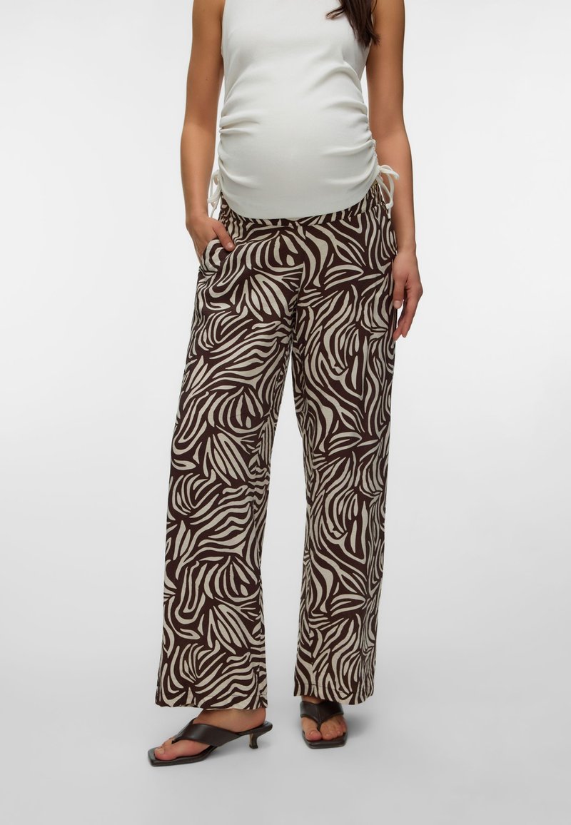 Femme enceinte portant un top blanc sans manches froncé, un pantalon large imprimé zèbre marron et beige, et des sandales à talons noires avec boucle pour les orteils.