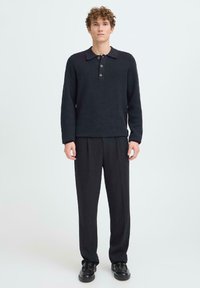 Sötétkék kötött pulóver hosszú ujjakkal és gallérral, három gombos elülső részszel, laza fekete nadrággal és fekete cipővel párosítva.