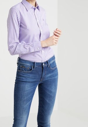 Femme portant un jean bleu slim et une chemise à manches longues à rayures verticales lavande et blanches, posant avec les mains levées près de la poitrine.