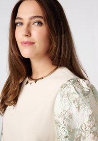 Haut beige avec des manches puff texturées au motif vert pastel. Présente un collier en or avec des accents en forme de cœur. Peau lisse et cheveux raides.