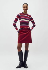Maglione a coste rigato in viola, rosso e crema; gonna mini in corduroy rossa con bottoni laterali; stivali neri in pelle al ginocchio.